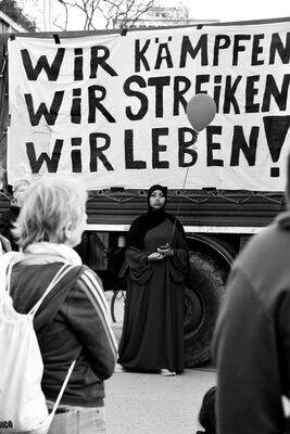 Black and white photograph of a woman in dark clothing speaking at a demonstration, beneath a hand-painted banner reading “Wir kämpfen, wir streiken, wir leben!”