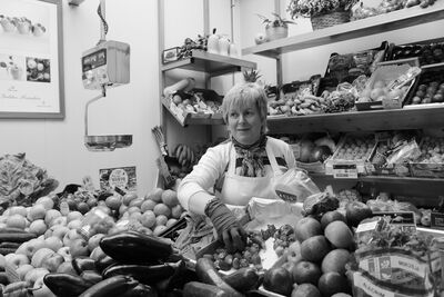 Black and white photograph of a smiling market vendor in an apron behind a stall piled with aubergines, peppers and other vegetables