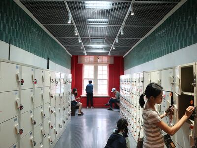 Visitors collecting their belongings from rows of cream lockers in a museum cloakroom corridor with teal tiled walls